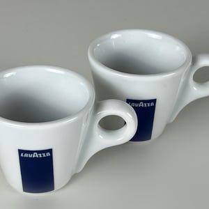 Pode incluir: Duas chávenas de café expresso de cerâmica branca com pegas curvas. Cada chávena apresenta um retângulo azul com o logótipo "LAVAZZA". As chávenas estão sobre um fundo cinzento claro.