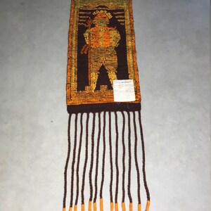 May include: A brown and orange woven wall hanging with a fringe. The hanging depicts a stylized figure in a brown, orange, and yellow color scheme.