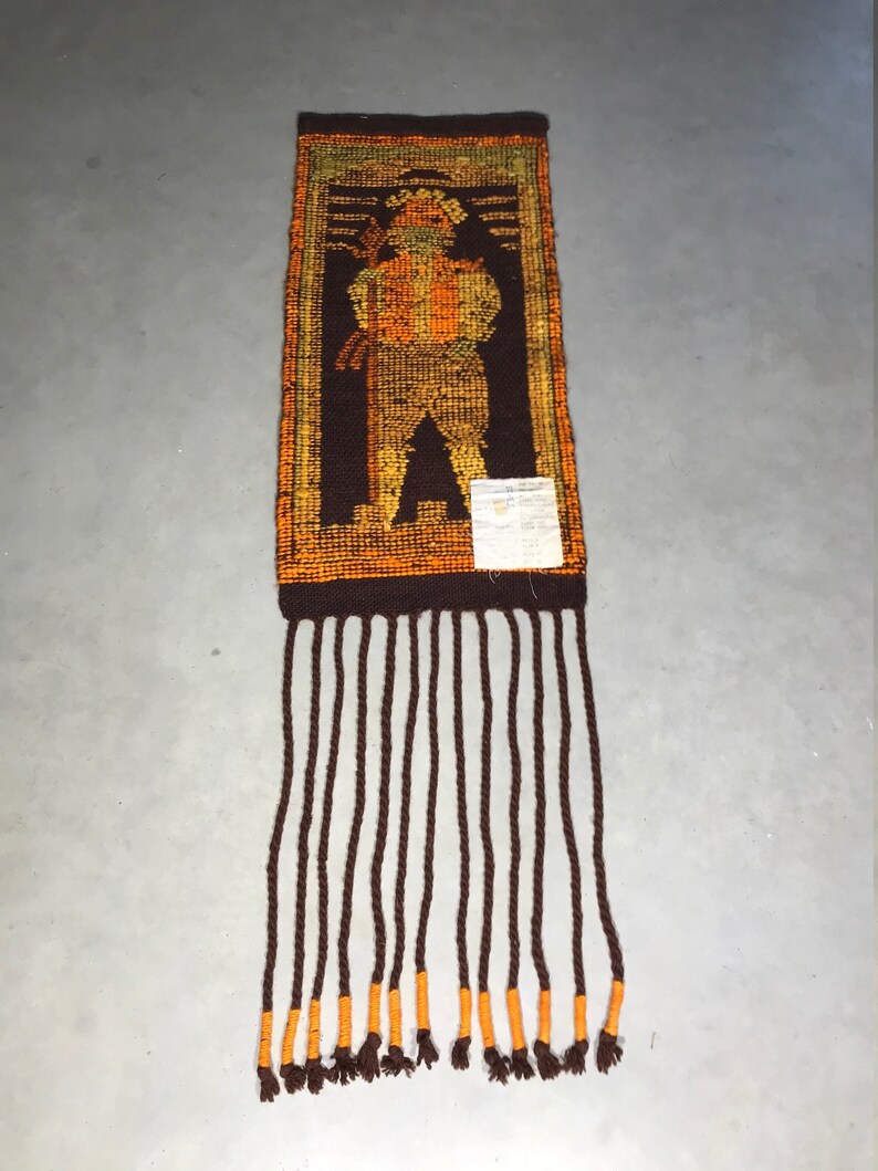 May include: A brown and orange woven wall hanging with a fringe. The hanging depicts a stylized figure in a brown, orange, and yellow color scheme.