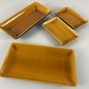 Op de afbeelding: Een set van vier rechthoekige serveerschalen in verschillende maten. De schalen hebben een glanzend, mosterdgeel interieur en een donkerbruine rand. De schalen zijn gerangschikt op een lichtgrijze ondergrond, waarbij hun verschillende afmetingen en vormen worden getoond.