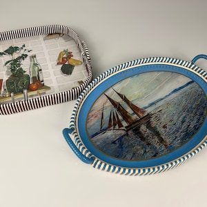 Op de afbeelding: Twee geweven dienbladen, een met een bruin-wit gestreept patroon en een keukenscène, de andere met een blauw-wit gestreept patroon en een zeilboot op zee.