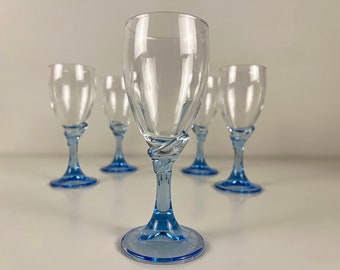 Puede incluir: Conjunto de cinco copas de vino de vidrio transparente con tallos y bases azules. Las copas están dispuestas en fila sobre una superficie blanca.