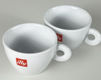 May include: Two white ceramic espresso cups with rounded handles. Each cup has a red square logo with the word "illy" printed in white. The cups are on a light gray surface.