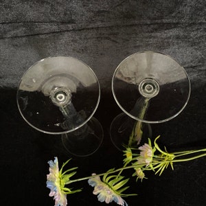 May include: Two clear glass candle holders with a tall, slender stem and a wide, round base. The candle holders are empty and are sitting on a black surface.