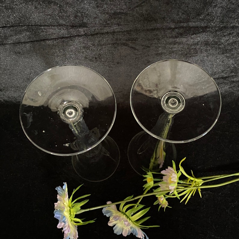 May include: Two clear glass candle holders with a tall, slender stem and a wide, round base. The candle holders are empty and are sitting on a black surface.