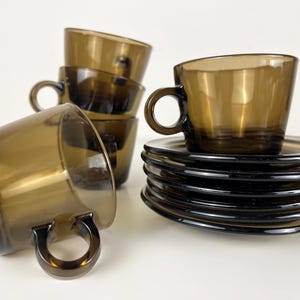 K&ouml;nnte beinhalten: Ein Set aus braunen Glas-Teetassen und Untertassen. Die Tassen haben eine abgerundete Form und einen Henkel. Die Untertassen sind rund und haben einen leicht erh&ouml;hten Rand.