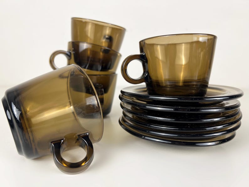 K&ouml;nnte beinhalten: Ein Set aus braunen Glas-Teetassen und Untertassen. Die Tassen haben eine abgerundete Form und einen Henkel. Die Untertassen sind rund und haben einen leicht erh&ouml;hten Rand.