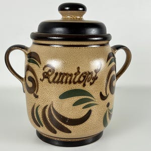 Könnte beinhalten: Beige Keramiktopf mit Deckel und zwei Griffen. Der Topf hat braune und grüne dekorative Akzente und das Wort "Rumtopf" in brauner Schrift. Der Deckel und der Boden sind dunkelbraun.