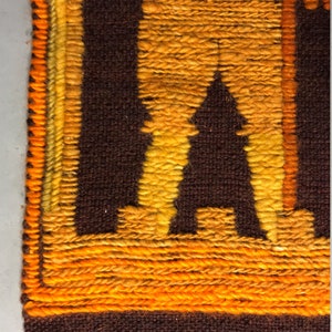 May include: A brown and orange woven wall hanging with a geometric design. The design features a figure with a long, flowing robe and a staff. The hanging has a fringe border.