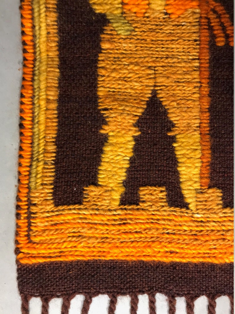 May include: A brown and orange woven wall hanging with a geometric design. The design features a figure with a long, flowing robe and a staff. The hanging has a fringe border.