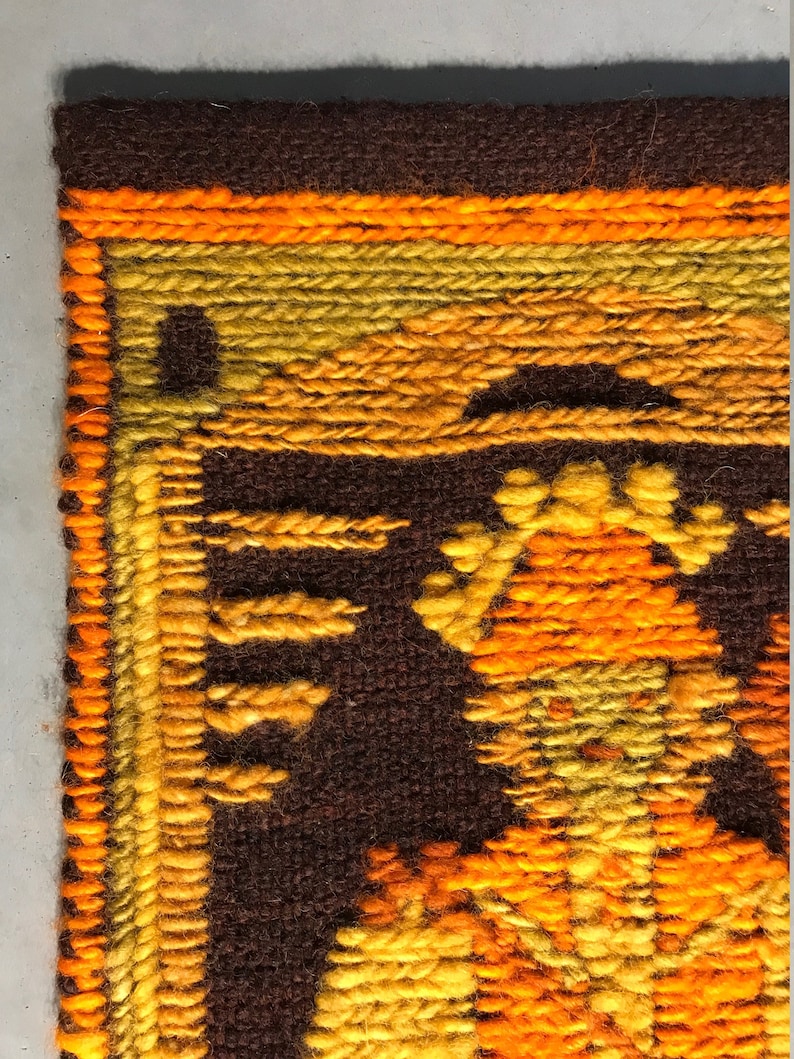 May include: A close-up of a brown, orange, and yellow woven wall hanging with a geometric pattern. The hanging is made of yarn and has a fringe border.