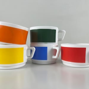 Op de afbeelding: Set van vier witte keramische mokken met gekleurde banden. De mokken hebben een groene, blauwe, gele en rode band.