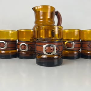 Op de afbeelding: Set van zes amber glazen met bruine leren banden en zilveren gespen. De glazen zijn in een rij geplaatst met een bijpassende amber glazen kan in de achtergrond.