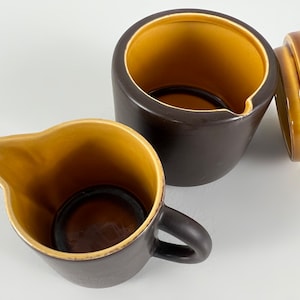 May include: A brown ceramic creamer and sugar bowl set with a contrasting yellow-brown interior. The creamer has a handle and spout, while the sugar bowl has a lid. The set is likely for serving coffee or tea.
