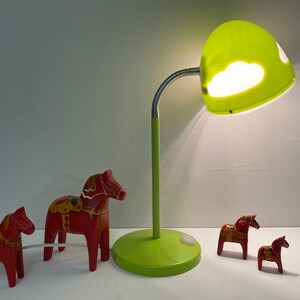 May include: Four red wooden toy horses, also known as Dala horses, with painted details, are arranged on a white surface. A green desk lamp with a cloud-shaped shade illuminates the scene.