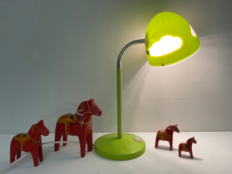 May include: Four red wooden toy horses, also known as Dala horses, with painted details, are arranged on a white surface. A green desk lamp with a cloud-shaped shade illuminates the scene.