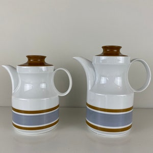 Op de afbeelding: Twee witte keramische theepotten met bruine deksels en blauwe en bruine strepen. De grotere theepot staat rechts en de kleinere links.