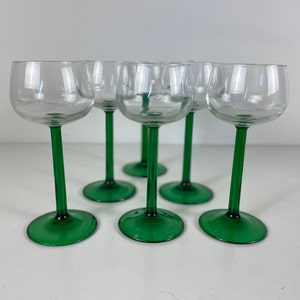 Op de afbeelding: Set van zes heldere glazen wijnglazen met groene stelen en voetjes. De glazen zijn in een halve cirkel op een wit oppervlak gerangschikt.