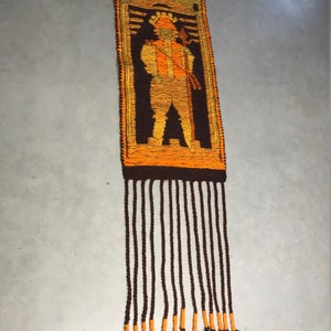 May include: A brown and orange woven wall hanging with a fringe. The hanging features a stylized figure in a brown, orange, and yellow color scheme.