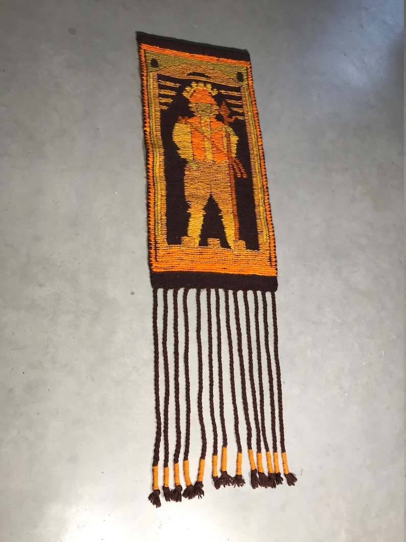 May include: A brown and orange woven wall hanging with a fringe. The hanging features a stylized figure in a brown, orange, and yellow color scheme.