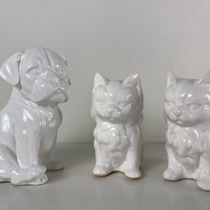Könnte beinhalten: Drei weiße Keramikfiguren eines Hundes und zweier Katzen. Der Hund sitzt mit dem Kopf zur Seite geneigt. Die Katzen sitzen beide mit dem Schwanz um die Beine gewickelt.