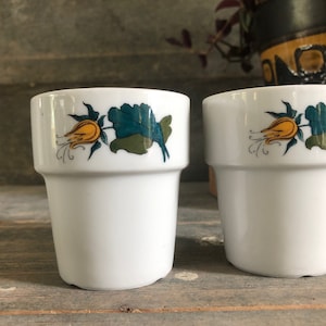 May include: Two white ceramic cups with a floral design. The cups have a yellow flower with green leaves and a white background.