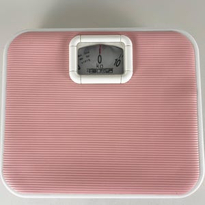 May include: A pink and white bathroom scale with a ridged surface. The scale has a white framed display showing the weight in kilograms. The scale reads zero. The brand name is visible on the display.
