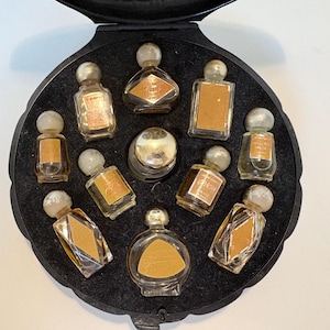 Op de afbeelding: Een vintage parfumset met een zwarte behuizing en een cirkelvormig interieur. De behuizing bevat elf kleine glazen parfumflesjes in verschillende vormen, elk met een zilveren dop en een gouden etiket. De set wordt gepresenteerd op een wit oppervlak.