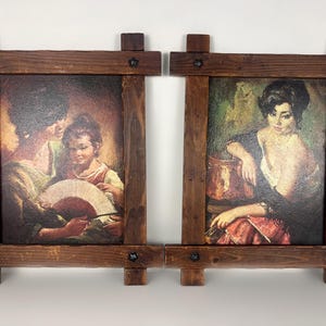 Può includere: Due stampe vintage incorniciate. Ogni stampa è in una cornice di legno marrone scuro con un design rustico. Una stampa raffigura una donna e un bambino con un ventaglio, l'altra una donna con una pentola di rame. L'opera d'arte ha una calda tavolozza di colori.