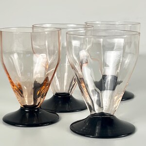 Op de afbeelding: Vier vintage dessertglazen van helder glas met een roze tint en zwarte voetjes. De glazen hebben een uniek, facet geslepen ontwerp en een uitlopende vorm, perfect voor het serveren van ijs of andere lekkernijen.