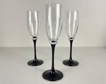 May include: Three clear glass champagne flutes with black stems. The flutes are arranged in a row on a white surface.