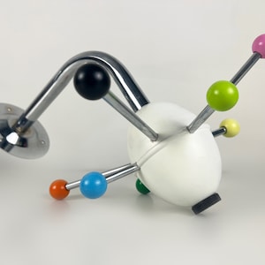 May include: A unique, abstract wall-mounted coat rack. It features a white, egg-shaped central form with multiple metal rods extending outwards, each tipped with a different colored sphere. The rack also has a curved, chrome-finished arm and a circular mounting plate.