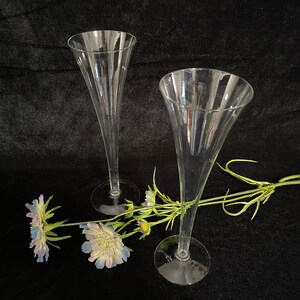 May include: Two clear glass champagne flutes with a delicate stem and a wide bowl. The flutes are arranged on a black surface with a sprig of blue and white flowers.