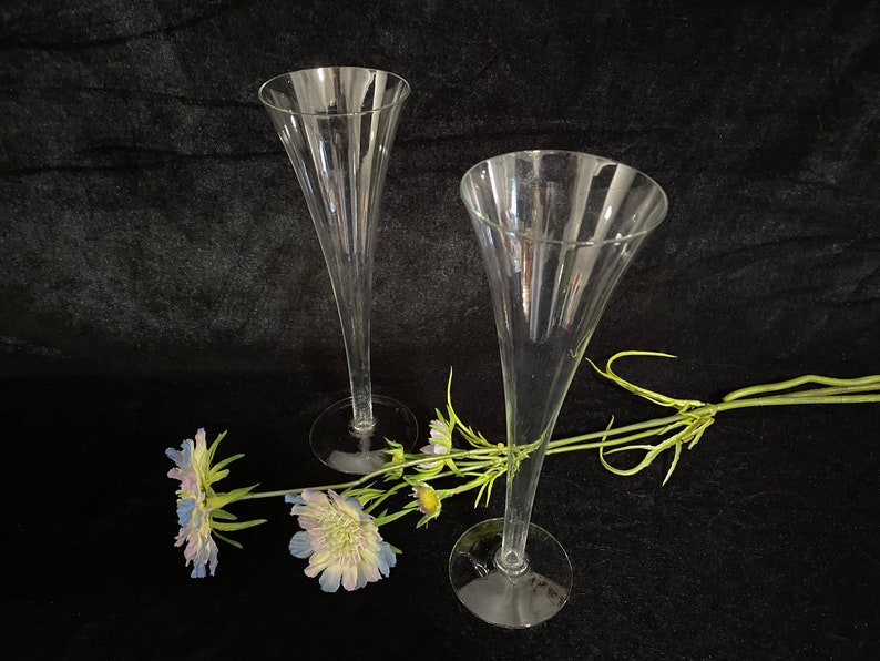 May include: Two clear glass champagne flutes with a delicate stem and a wide bowl. The flutes are arranged on a black surface with a sprig of blue and white flowers.
