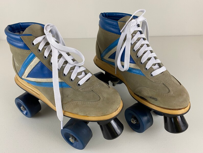 May include: A pair of vintage roller skates with gray suede uppers, blue and white accents, and white laces. The skates have blue wheels and black trucks.