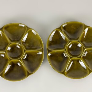 May include: Two olive green ceramic oyster plates. Each plate has a central well and six surrounding compartments. The plates have a glossy finish and are set against a white background. The plates are designed for serving oysters.
