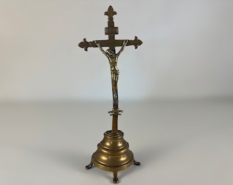 May include: A gold-toned metal crucifix with a figure of Jesus Christ on the cross. The crucifix is mounted on a decorative stand with a round base and three legs.