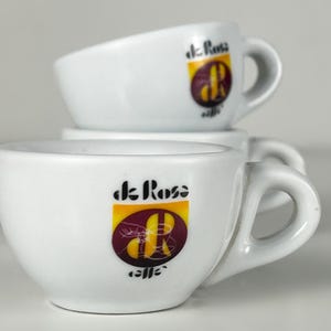 Puede incluir: Tres tazas de espresso blancas apiladas. Cada taza tiene un asa y un logotipo que dice "de Rose caffe" en texto negro con un emblema amarillo y granate. Para café.