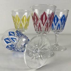 Puede incluir: Juego de cuatro copas de vino transparentes con diseños geométricos coloridos. Una copa está tumbada de lado. Los diseños son amarillos, rojos y azules. Las copas tienen un patrón de estrella en la base.