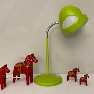 May include: Four red wooden Dala horses, a traditional Swedish folk art, are arranged on a table with a green desk lamp with a white cloud design on the shade.