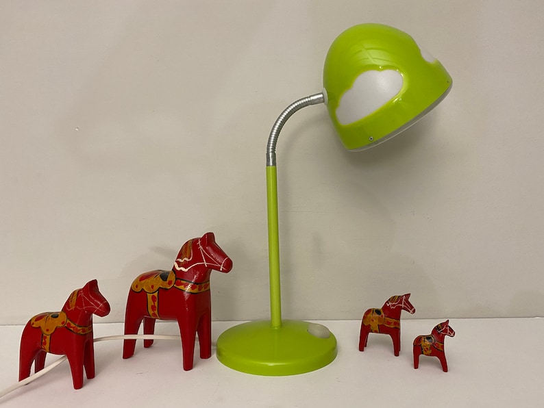 May include: Four red wooden Dala horses, a traditional Swedish folk art, are arranged on a table with a green desk lamp with a white cloud design on the shade.
