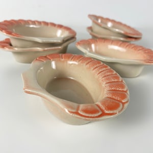 May include: Set of six small, oval-shaped ceramic dishes with a light beige glaze and a scalloped, peach-coloured rim.