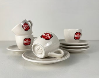 May include: A set of white espresso cups and saucers. Each cup has a handle and a red and white logo that says "cibo caffe". The saucers are round and white. The cups are stacked and arranged on the saucers.