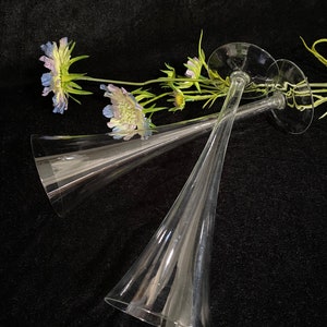 May include: Two clear glass champagne flutes with a wide base and a long, narrow stem. The flutes are lying on a black surface with a sprig of purple and green flowers.