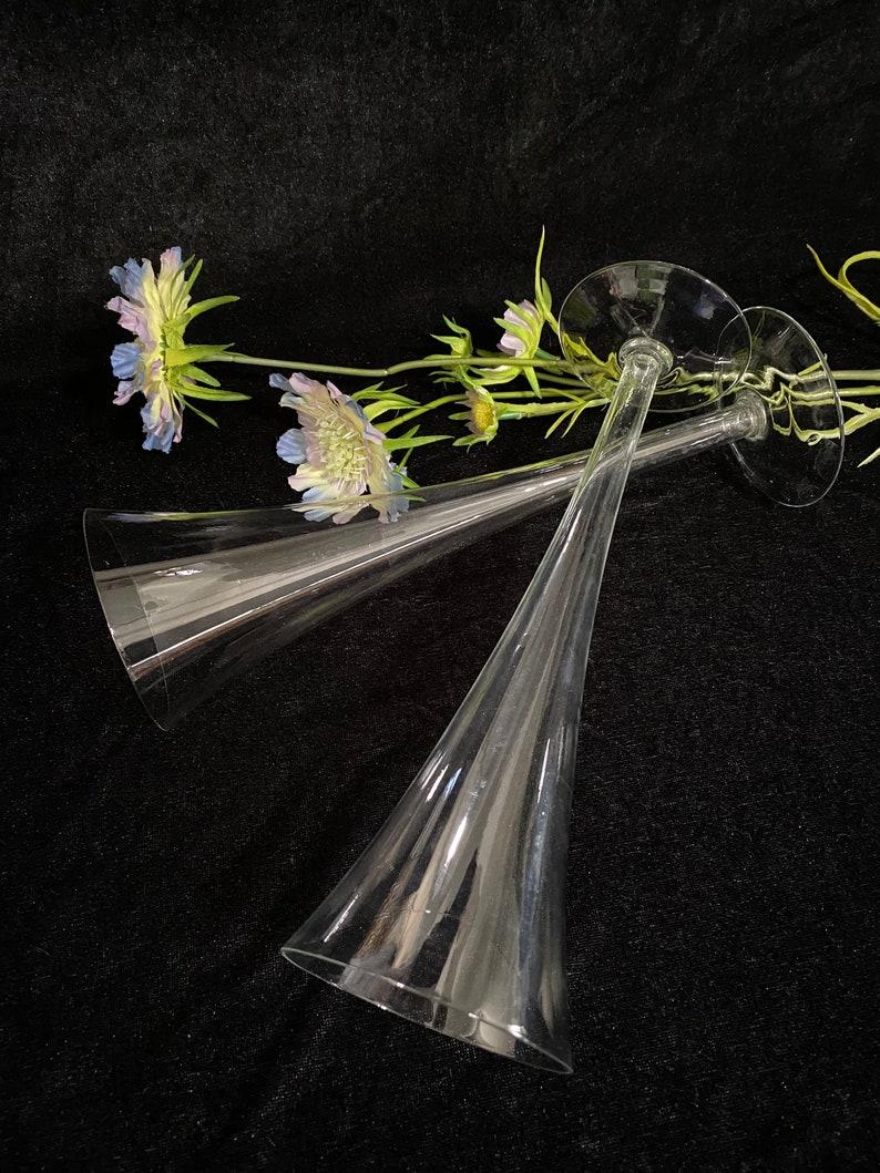 May include: Two clear glass champagne flutes with a wide base and a long, narrow stem. The flutes are lying on a black surface with a sprig of purple and green flowers.