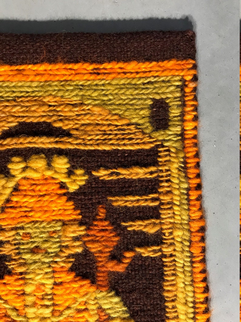 May include: Close-up of a brown, orange, and yellow woven wall hanging with a geometric pattern. The hanging has a fringe border.
