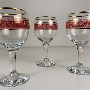 May include: Three clear glass wine glasses with gold rims and a red and white patterned band around the middle.