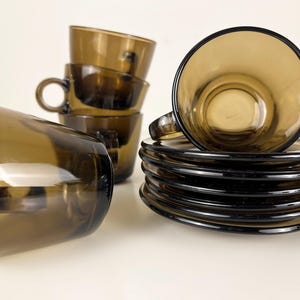 K&ouml;nnte beinhalten: Set aus braunen Glas-Teetassen und Untertassen. Die Tassen und Untertassen sind gestapelt und auf einer wei&szlig;en Oberfl&auml;che angeordnet.