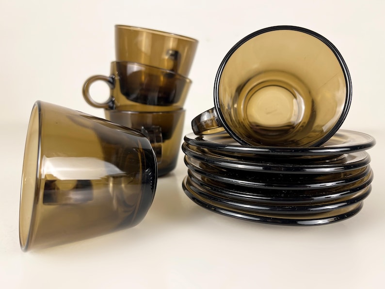 K&ouml;nnte beinhalten: Set aus braunen Glas-Teetassen und Untertassen. Die Tassen und Untertassen sind gestapelt und auf einer wei&szlig;en Oberfl&auml;che angeordnet.