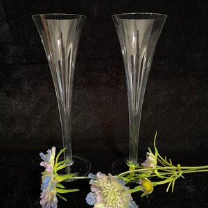 May include: Two clear glass champagne flutes with a fluted design. The flutes are on a black surface with blue and green flowers.
